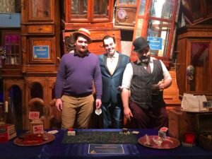 Adam Stone Magic Three Men Stand Together Behind a Casino style Poker Table in a Wood paneled Room Posing for a Photo the Man on the Left Wears a Purple Sweater and a Straw Hat the Middle Man Wears a Blue Suit and the Man on the Right Wears a Vest and Bowler Hat with a Beard Holding a Mug of Drinks