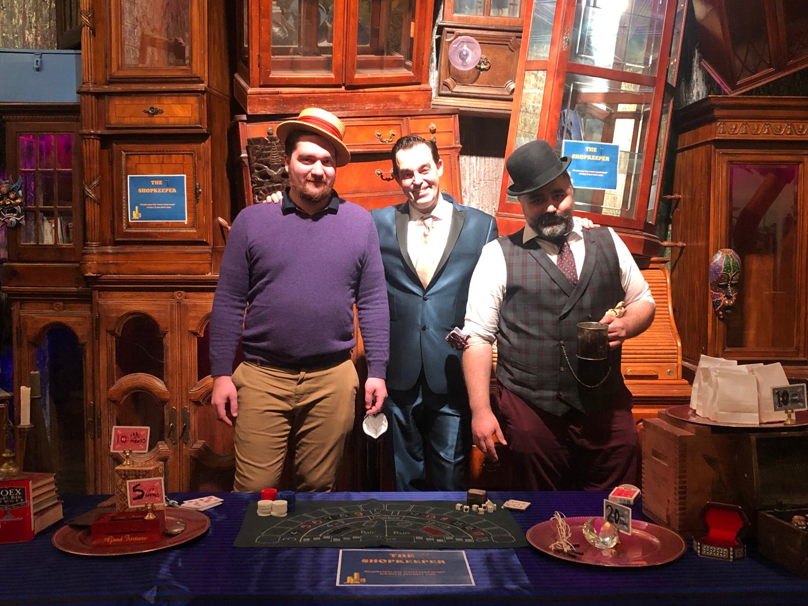 Adam Stone Magic Adam Stone Magic Three Men Stand Together Behind a Casino Style Poker Table in a Wood Paneled Room Posing for a Photo the Man on the Left Wears a Purple Sweater and a Straw Hat the Middle Man Wears a Blue Suit and the Man on the Right Wears a Vest and Bowler Hat with a Beard Holding a Mug of Drinks