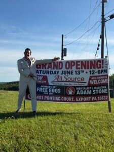 Mentalist Adam Stone in a Suit Standing Beside a Grand Opening Sign for All Source Featuring His Name As the Featured Mentalist and Magician