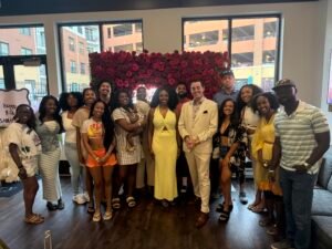 Group of People Smiling and Posing in Front of a Dense Red Rose Backdrop Indoors Wearing Summer Outfits